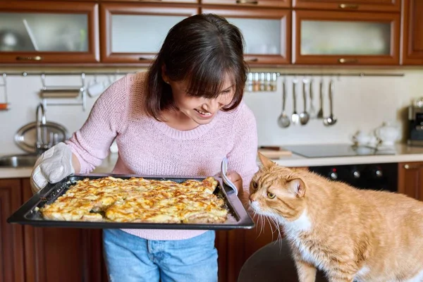 Woman with a baking sheet of baked meat and a funny ginger domestic cat asking for meat