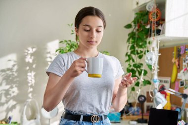 14-15 yaşlarında elinde fincanla güneşli pencerenin yanında duran bir kız. Güzel, çekici bir kadın evde dinleniyor, gülümsüyor. Ergenlik, yaşam tarzı, gençlik kavramı.