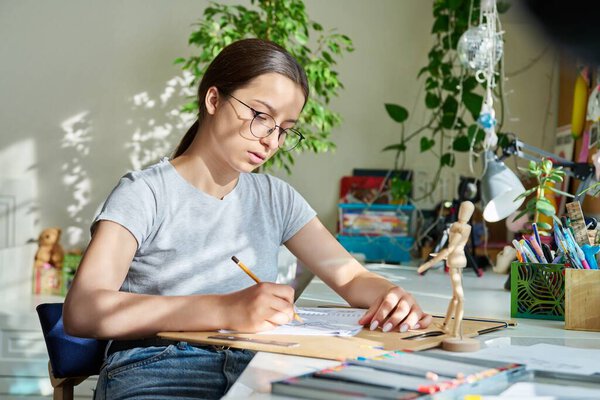 Teenage creative girl artist drawing with a pencil, sitting at the table at home, using wooden mannequin. Creativity, school, hobby, leisure, lifestyle, adolescence, youth concept