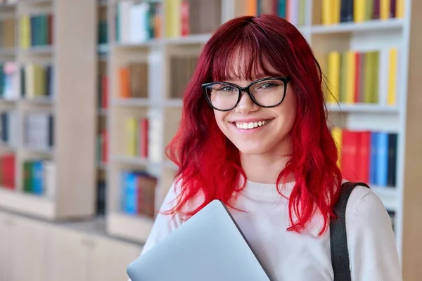 Portrait of female student with laptop backpack, looking at camera in library. Beautiful fashionable girl with dyed red hair wearing glasses, smiling with teeth. College, university, education, youth