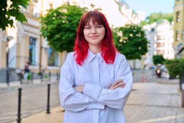 Kollarını kavuşturup kameraya bakan kendine güvenen genç bir bayan. Şehirde kızıl boyalı saçlı, güzel, modaya uygun bir kız. Güzellik, gençlik, yaşam tarzı konsepti