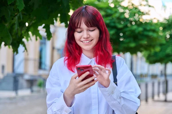 Beautiful smiling teenage girl looking at phone on city street. Female teenager with red dyed hair with backpack using smartphone. Lifestyle, technology, communication, youth, young people