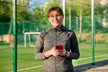 Portrait of smiling middle aged woman looking at camera at outdoor stadium. Active female in wireless headphones sportswear with smartphone in hand near football field. Urban sport, healthy lifestyle clipart