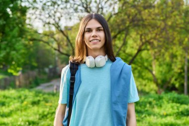 Sırt çantalı, yeşil park kampüs geçmişine bakan yakışıklı bir öğrencinin açık hava portresi. Uzun saçlı, gülümseyen genç bir erkek. Gençlik, eğitim, gençlik kavramı