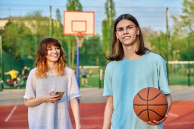 Portrait of young guy playing basketball on an outdoor game court, girl is out of focus. Handsome teenager smiling looking at camera with ball in hands in sunset light. Youth, active healthy lifestyle