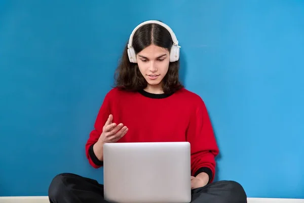 十几岁的男生在耳机中使用笔记本电脑进行会议视频通话 坐在地板上蓝色背景 电子教育 在线学习 技术概念 — 图库照片
