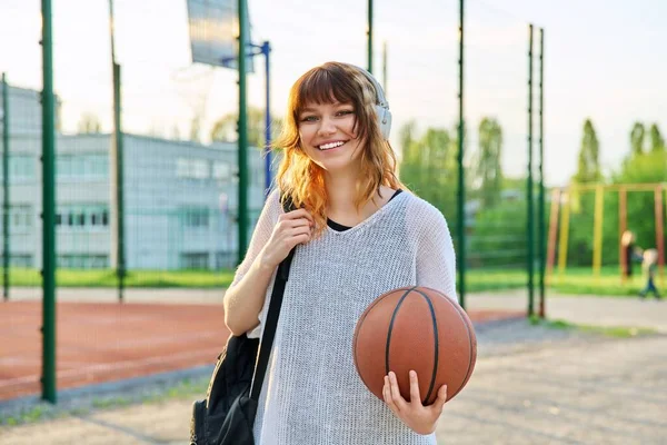 バックパックを持つヘッドフォンで美しい赤毛の十代の女性学生は カメラバスケットボールの屋外コートを見て笑顔ボールを保持します アクティブな健康的なライフスタイル 都市スポーツ — ストック写真