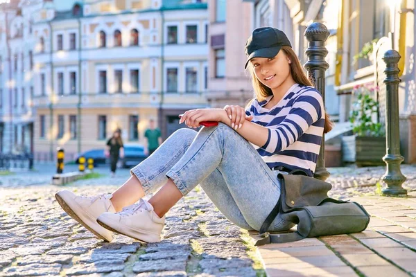 漂亮的少女坐在人行道上看着镜头 在城市街道上戴着背包头戴帽子的时髦女生 生活方式 青年概念 — 图库照片