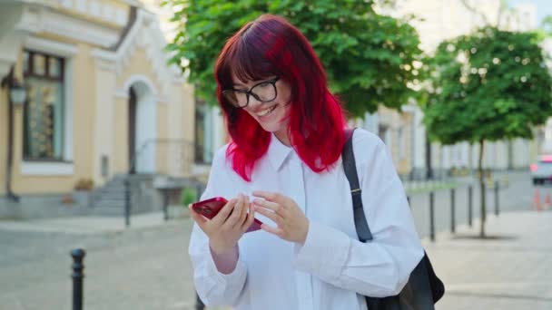Teenager modische positive Studentin Blick auf Smartphone-Bildschirm, Mädchen lächelnd Nachricht beobachten. Frau mit Brille und knallrot gefärbten Haaren auf der Straße der Stadt, Jugend-Lifestyletechnologie
