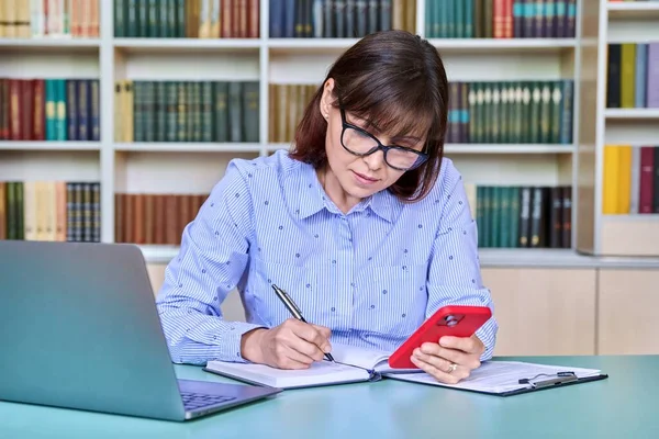 Middle-aged female teacher working in library, using laptop, smartphone. Education, knowledge, school, college, university technology teaching concept