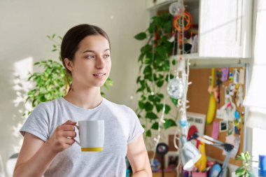 14-15 yaşlarında elinde fincanla güneşli pencerenin yanında duran bir kız. Güzel, çekici bir kadın evde dinleniyor, gülümsüyor. Ergenlik, yaşam tarzı, gençlik kavramı.