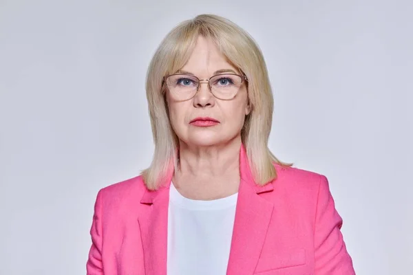 Portrait of senior business successful woman in glasses looking at camera, on light studio background. Confident beautiful blonde female in pink jacket. Confidence professionalism business, 50s people