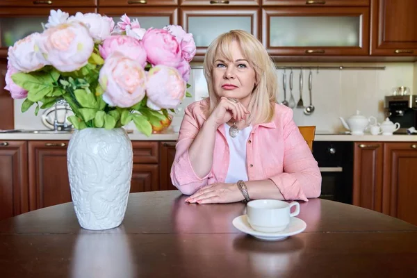 Beautiful mature woman looking at camera sitting at table at home with bouquet of flowers in vase, cup of coffee. Beauty senior female, 50s age, confidence, success, health, joy, happiness concept