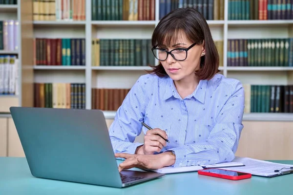 Middle-aged female teacher working in library, using laptop writing in papers at her desk. Education, knowledge, school, college, university, technology, teaching concept