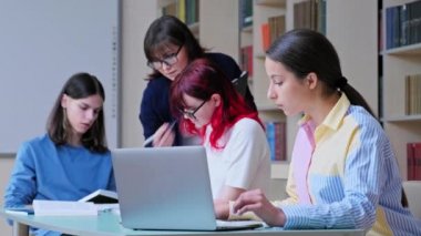 Bir grup genç öğrenci okul kütüphanesi sınıfında ders çalışıyor. Genç kız dizüstü bilgisayar kullanıyor, öğretmen öğrencilere yardım ediyor. Lise, öğrenim, eğitim, bilgi, ergenlik