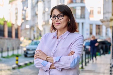 Dışarıdaki kameraya bakan kendinden emin orta yaşlı bir kadının portresi. Gözlüklü, gülümseyen bir kadın, kollarını şehir caddesinde kavuşturmuş. İş, yaş, portre, güven, 40 'lı yılların insan kavramı