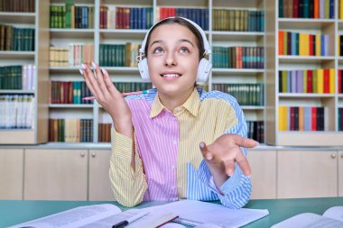 Kulaklıklı genç bir kız web kamerasına bakıyor, online ders sohbeti danışmanlığı yapıyor. Çalışmak için video konferansı kullanan genç bir kız öğrenci, sınıf kütüphanesi geçmişi. E-öğrenme, ergenlik