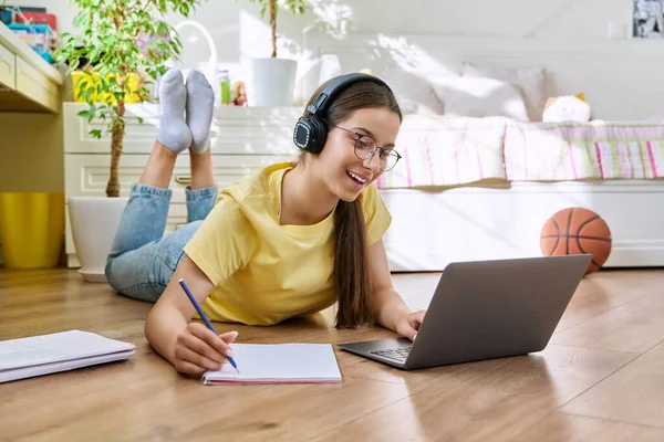 Teenage girl with glasses studying at home using laptop. Female with headphones and textbooks lying on floor in room. Education, adolescence, high school concept