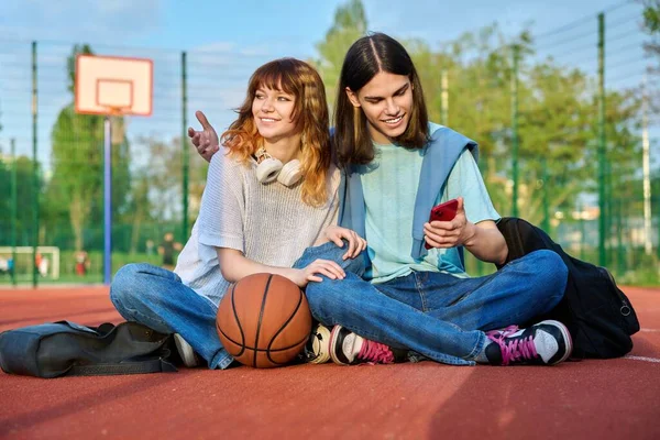 10代の学生の友人のカップルは 屋外のバスケットボールコートに座って ボールのバックパックで 笑って 話して スマートフォンで探している アクティブな健康的なライフスタイル 都市スポーツの概念 — ストック写真