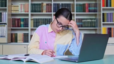 Gözlüklü liseli kız kütüphanede kitap ve dizüstü bilgisayar kullanarak ders çalışıyor. Teknoloji, eğitim, bilgi, ergenlik kavramı