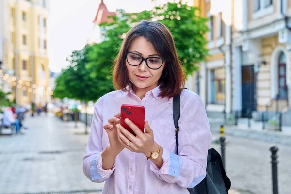 Middle aged smiling woman looking into smartphone on city street. Beautiful mature female using mobile online applications. Technology, business, lifestyle, urban style, 40s age people concept