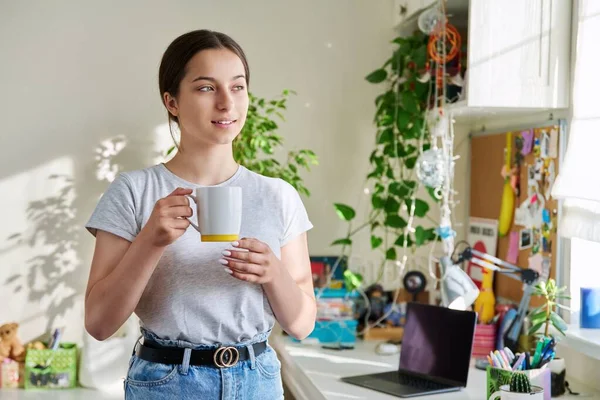 14-15 yaşlarında elinde fincanla güneşli pencerenin yanında duran bir kız. Güzel, çekici bir kadın evde dinleniyor, gülümsüyor. Ergenlik, yaşam tarzı, gençlik kavramı.