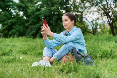 Genç kız öğrenci akıllı telefon kullanarak çimlerin üzerinde dinleniyor. Bahçede basketbol topu ve sırt çantasıyla çekici bir genç kız. Lise, teknoloji, aktif sağlıklı yaşam tarzı, ergenlik kavramı.