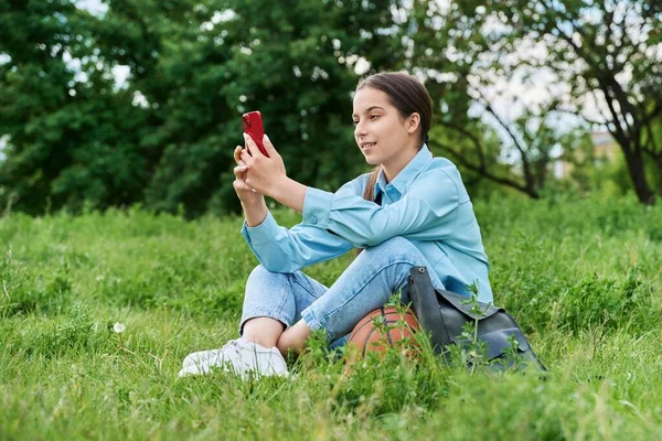 十几岁的女生坐在草地上 用智能手机休息 迷人的少女在草坪上打篮球 积极健康的生活方式 青春期观念 — 图库照片