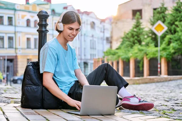 Teenage male student in headphones using laptop outdoor, on city street. Guy 17, 18 years old looking at screen, sitting on sidewalk. College, high school, youth, education, technology concept