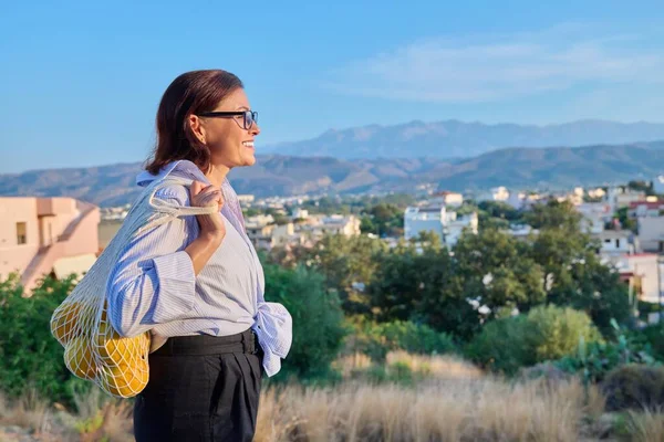 Happy calm woman enjoying vacation in nature, profile view, sky city in copy space mountains. Beautiful mature female with bag of organic oranges. Summer, scenic landscape, tourism, people concept