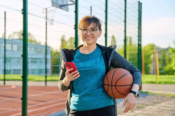 Active mature woman with basketball ball and smartphone, outdoor basketball court background. Middle-aged female in sportswear wearing wireless headphones. Sporty healthy lifestyle, leisure activity
