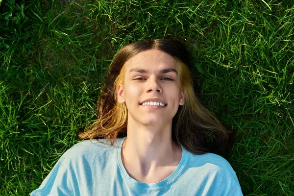 Smiling young guy looking at camera, lying on grass, green background. Handsome teenage male with long hair, trendy hairstyle in blue t-shirt. Youth, lifestyle, people concept