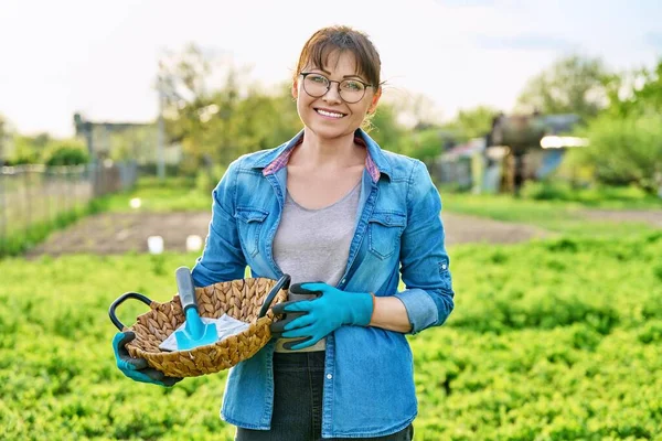 Middle-aged woman looking at camera in spring garden, in gloves with shovel and seeds in packs. Spring time, season to plant, farm, cottage, growing organic vegetables