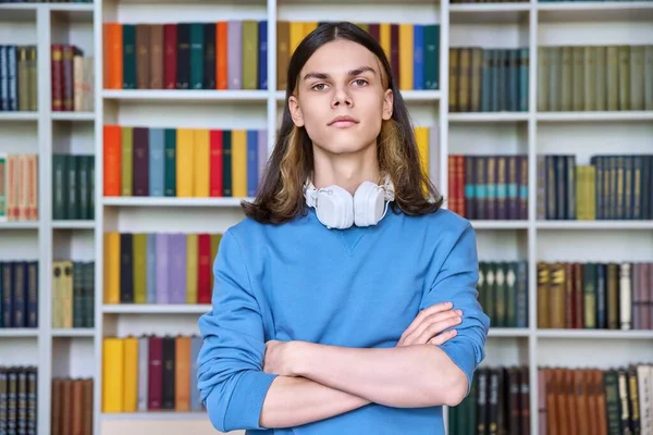 Portrait of serious guy student looking at camera in library. Confident handsome teenage male with headphones crossed arms. Education, youth,college, university concept