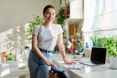 Genç kız kameraya bakıyor, evde duruyor ve masada dizüstü bilgisayarı var. Ergenlik, yaşam tarzı, öğrenci hayatı, gençlik kavramı.