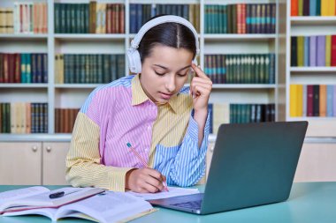Kulaklıklı liseli kız kütüphanede kitap ve dizüstü bilgisayar kullanarak ders çalışıyor. Teknoloji, eğitim, ergenlik kavramı