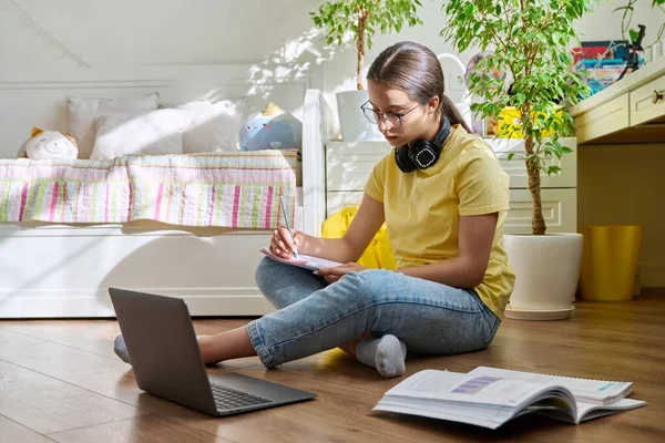 Teenage girl with glasses studying at home using a laptop. Female with headphones and textbooks sitting on the floor in the room. Education, adolescence, high school concept