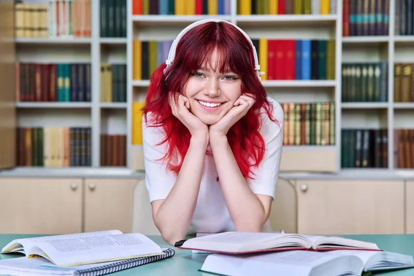 一个漂亮的女学生坐在图书馆的书桌前 看着相机的画像 时髦的红色染发耳机微笑的少女 教育知识 青春期 — 图库照片