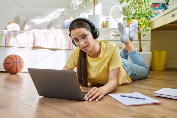Teenage girl with glasses studying at home using laptop. Female with headphones and textbooks lying on floor in room. Education, adolescence, high school concept