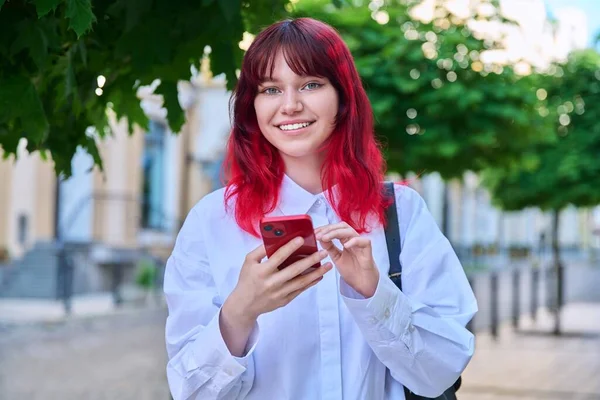 Outdoor portrait of fashionable teenage female student with smartphone in hands looking at camera, on city. Teenager with trendy colored hairstyle red hair. Youth fashion, beauty, technology lifestyle