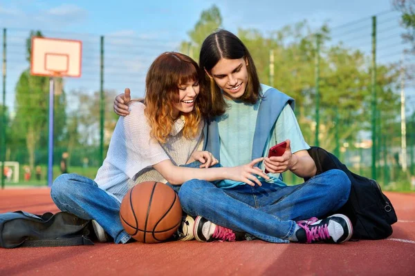 10代の学生の友人のカップルは 屋外のバスケットボールコートに座って ボールのバックパックで 笑って 話して スマートフォンで探している アクティブな健康的なライフスタイル 都市スポーツの概念 — ストック写真