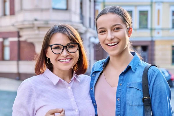 Portrait of happy mom and teenage daughter looking at camera outdoor on city street. Smiling mother and teen girl together. Family, two generations, lifestyle, leisure, people concept
