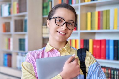 Kütüphanede kameraya bakan lise öğrencisinin portresi. Sırt çantası ve dizüstü bilgisayarıyla gözlüklü gülümseyen genç kız. Öğrenim, eğitim, bilgi, ergenlik kavramı