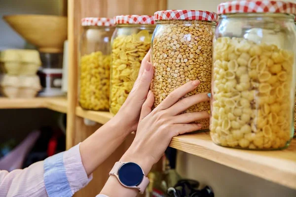 Storage of food in kitchen in pantry, womans hands with can of dry peas. Cereals, food in jars containers on wooden rack. Cooking at home, stocking food, household