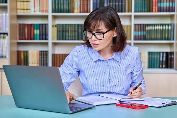 Middle-aged female teacher working in library, using laptop writing in papers at her desk. Education, knowledge, school, college, university, technology, teaching concept