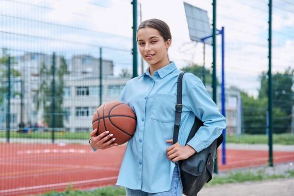 Portrait of teenage student girl with backpack and basketball ball, looking at camera, near basketball school outdoor court. Active healthy sport lifestyle, adolescence, school concept