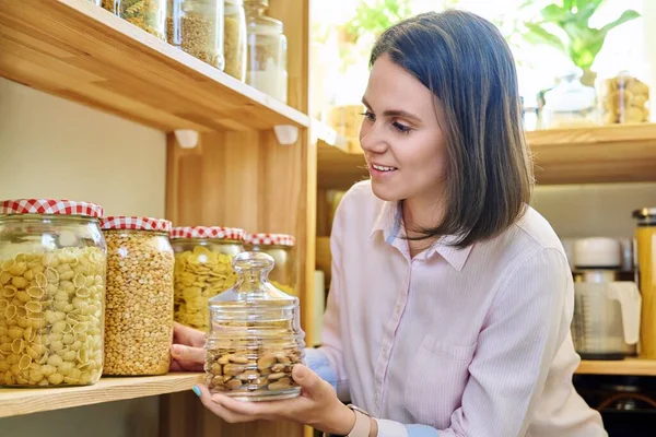 Young woman in kitchen with containers jars of nuts, cereals, pasta, dry fruits. Female in home pantry, food organization and storage, kitchen utensils, household, food preparation at home concept