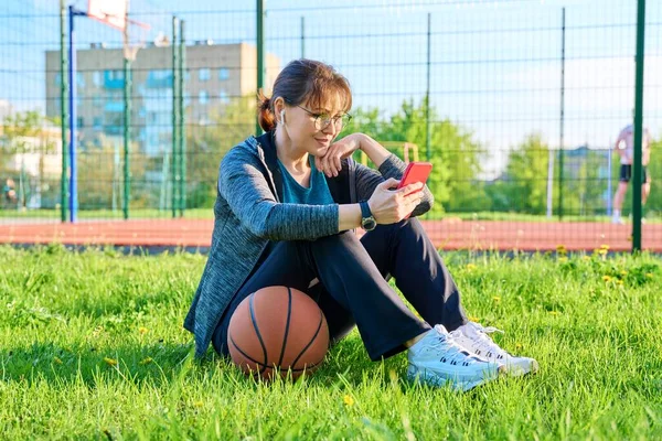 バスケットボールとスマートフォン 屋外バスケットボールコートの背景を持つアクティブな成熟した女性 草の上に座ってスポーツウェアのヘッドフォンで中年の女性 スポーツ健康的なライフスタイル レジャー活動 — ストック写真