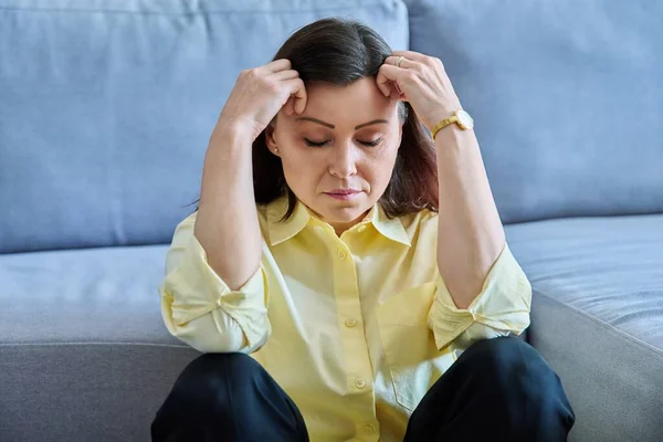 Sad upset middle-aged woman sitting on the floor near the sofa, unhappy female holds her head with her hands. Pain, stress, depression, mental health, mature age concept