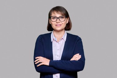 Gri soyutlanmış arka planda gülümseyen orta yaşlı bir iş kadını. 40 'lı yaşlarda kollarını kavuşturmuş kameraya bakan kendinden emin bir kadın. Öğretmen, psikolog, meslek, psikoloji terapisi, başarı, iş, personel
